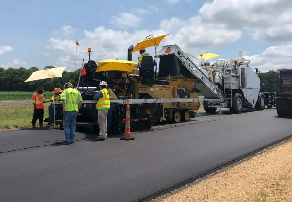 An image of construction workers using a paver machine to apply a pavement overlay to a roadway.